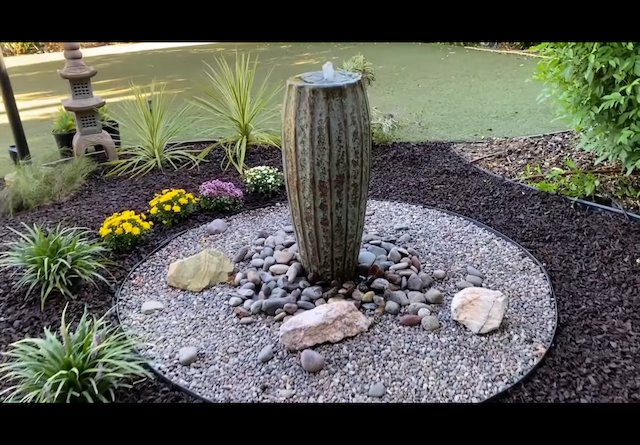 Tall ribbed ceramic bubbling fountain in a circular bed of river pebbles