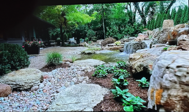 Wide stone waterfall spilling into a clear pond beside a patio and grill